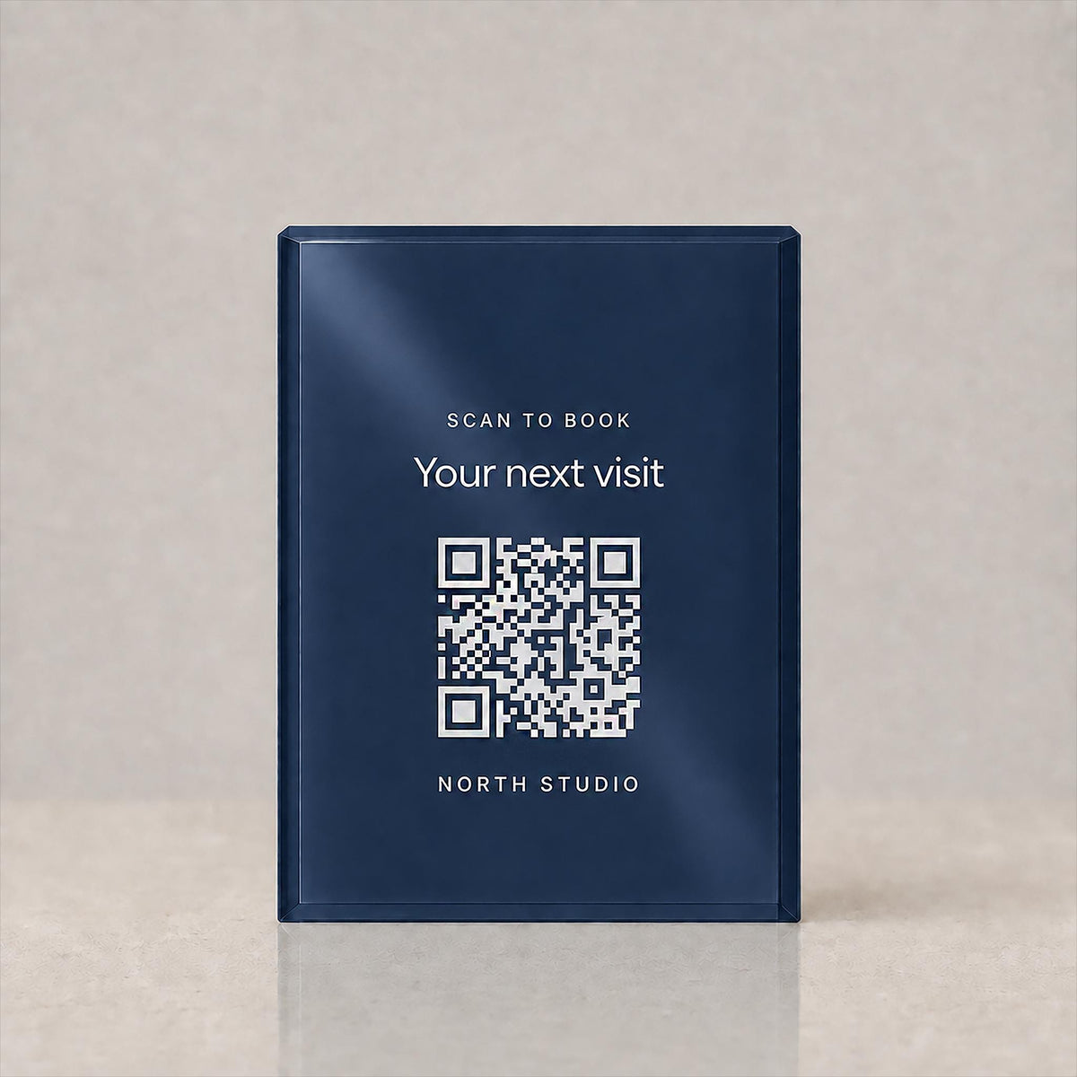 Front view of a navy acrylic QR code sign printed with Scan to book, Your next visit, a QR code and a studio name, on a plain background.