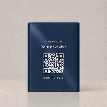Front view of a navy acrylic QR code sign printed with Scan to book, Your next visit, a QR code and a studio name, on a plain background.