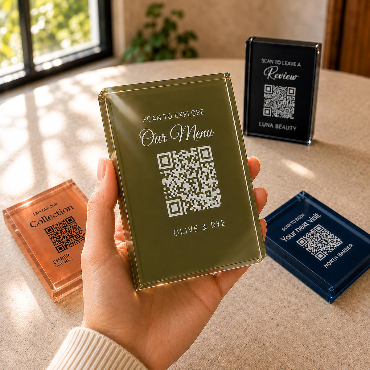 Hand holding an olive green acrylic QR code sign printed with a menu message, with terracotta, black and navy signs on the table behind.