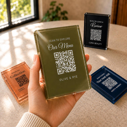 Hand holding an olive green acrylic QR code sign printed with a menu message, with terracotta, black and navy signs on the table behind.