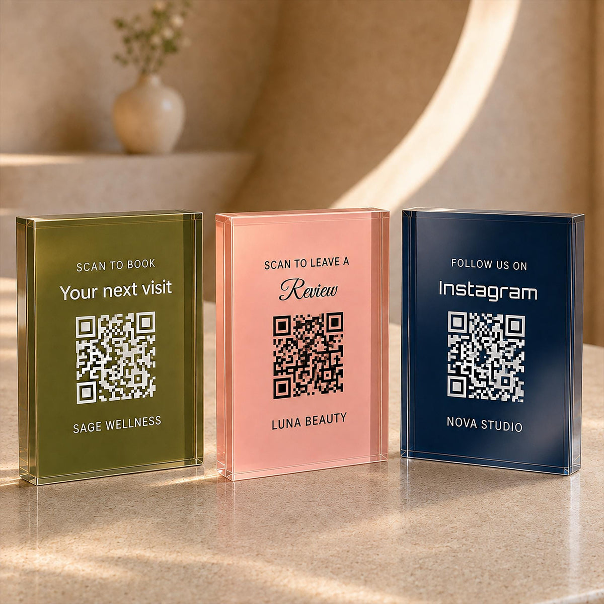 Three acrylic QR code signs in olive green, blush pink and navy, printed for booking, reviews and Instagram, standing on a stone counter.