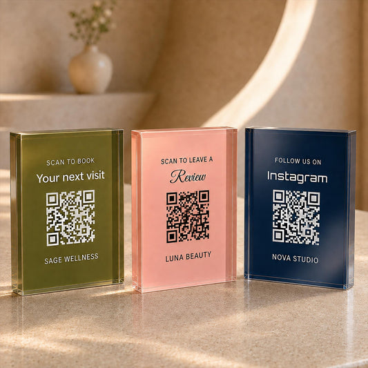 Three acrylic QR code signs in olive green, blush pink and navy, printed for booking, reviews and Instagram, standing on a stone counter.