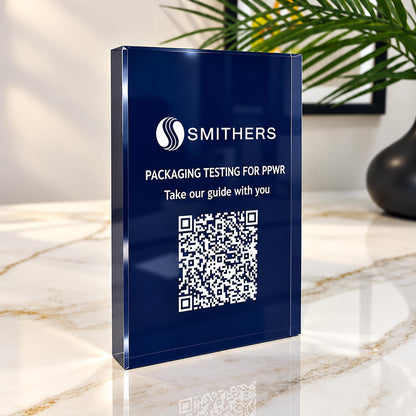 Navy acrylic QR code sign standing on a marble counter, printed with a company logo, a short message and a QR code, beside a plant.
